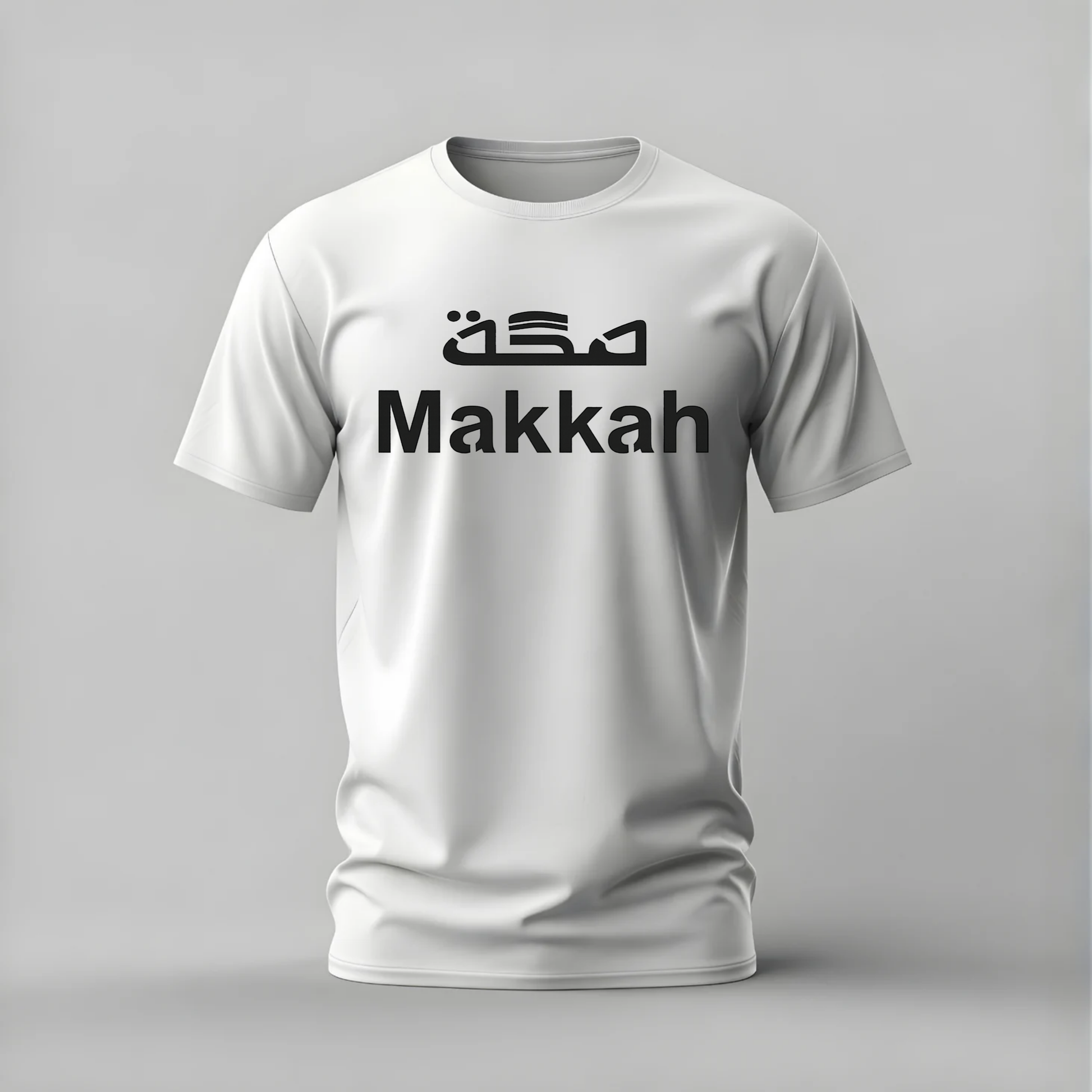 A white short-sleeve crewneck t-shirt with 'Makkah' printed in black across the chest, featuring stylized Arabic calligraphy above it, shown on a neutral gray background.